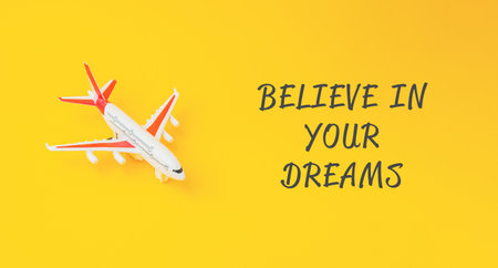 A small white airplane is on a yellow background with the words believe in your dreams written below itの写真素材
