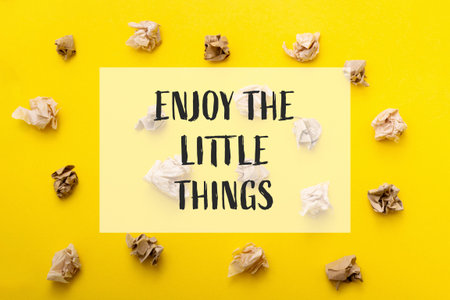 A yellow background with a pile of crumpled paper on it. The paper is scattered in various sizes and shapes, creating a sense of chaos and disorder. A white square with black text overlaying the background says, 'Enjoy the little things'.の写真素材