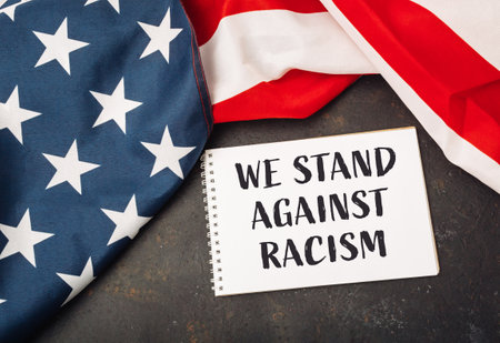 White piece of paper with the words We stand against racism written on it. The paper is placed on top of a red and white American flagの写真素材