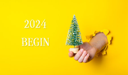 A hand holding a small Christmas tree in front of a yellow background with the words 2024 Begin written belowの写真素材