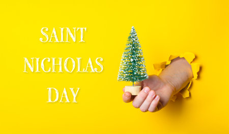 A hand holding a small Christmas tree in front of a yellow background with the words Saint Nicholas Day written belowの写真素材
