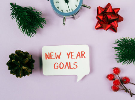 A clock sits on a table next to a white sign that says New Year Goals. The sign is surrounded by a bunch of green leaves and red berriesの写真素材