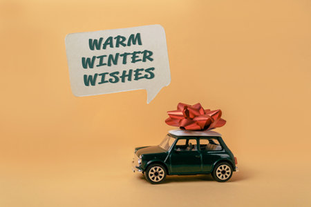 Green toy car with a red bow on top of it. The car is on a yellow background with the words warm winter wishes written above itの写真素材