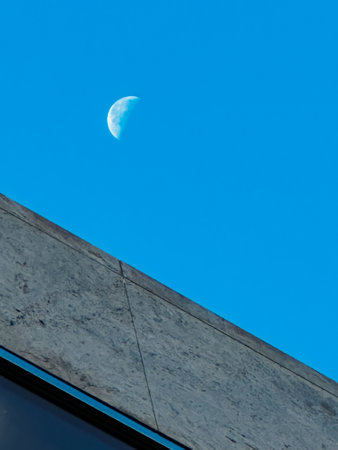 A crescent moon is in the sky above a building. The sky is clear and blue.の写真素材
