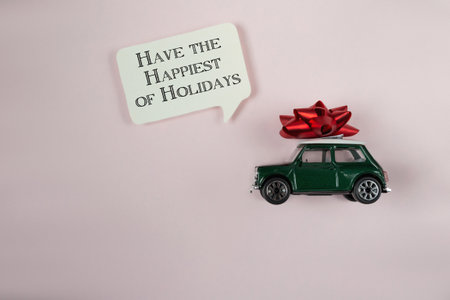 Green toy car with a red bow on top of it. The car is on a pink background with the words Have the happiest holidays written below itの写真素材