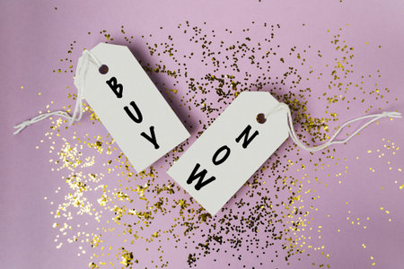 Two tags with the words buy and now are hanging from a string. The tags are on top of a glittery...