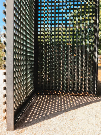 A black metal fence with a lattice design. The lattice is made of metal and has a diamond pattern. The fence is in a park and is surrounded by trees. The sun is shining on the fenceの写真素材