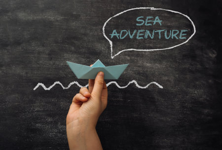A hand holding a paper boat in front of a chalkboard with the words Sea Adventure written in whiteの写真素材