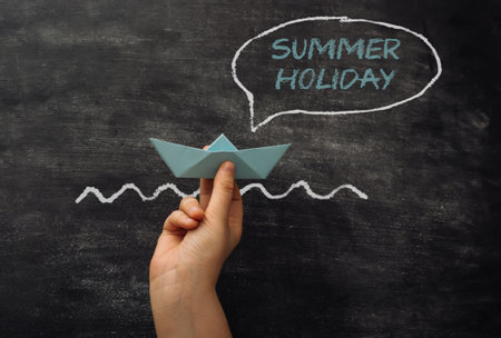 A hand holding a paper boat in front of a chalkboard with the words Summer Holiday written in whiteの写真素材