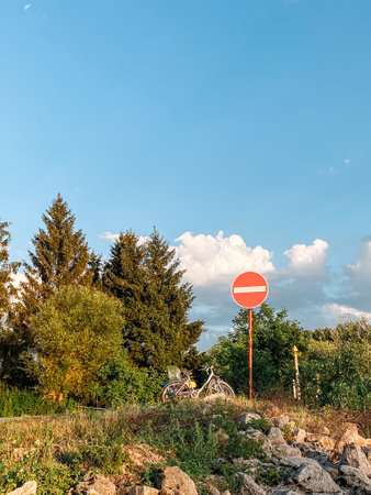 Red and white sign with a white line in the middle is on a hillside. The sign is surrounded by trees and a bicycle is parked nearby. The scene is peaceful and serene, with the blue sky aboveの写真素材
