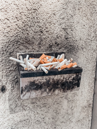 Pile of cigarette butts are in a container. The container is on a wall. The butts are scattered all over the containerの写真素材