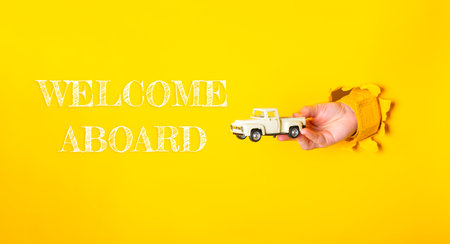 Hand holding a toy truck is holding a hole in a yellow background with the words welcome aboard written in whiteの写真素材