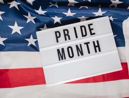 Pride month is written on a white sign that is placed on top of a red, white, and blue American flag