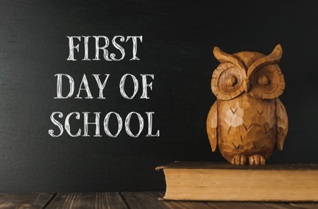 Wooden owl sits on a book next to a chalkboard that says First Day of School.の写真素材