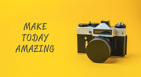 Camera is on a yellow background with the words Make today amazing written below it. The camera is black and silverの写真素材
