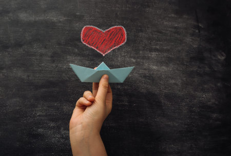 Hand holding a paper boat with a heart on it. Concept of love for sea travel or adventureの写真素材