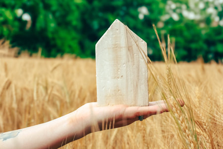 A hand presents a wooden house model amidst golden wheat, surrounded by lush greenery and a clear sky, symbolizing home and natureの写真素材