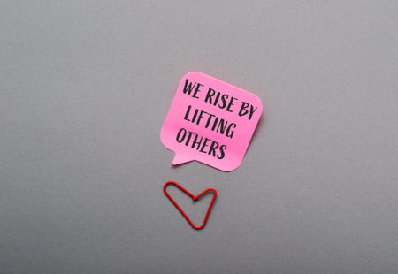 Pink and red sign that says We rise by lifting others is placed on a grey surface