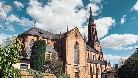 Historic stone church features intricate architecture with a tall spire, surrounded by lush greenery...