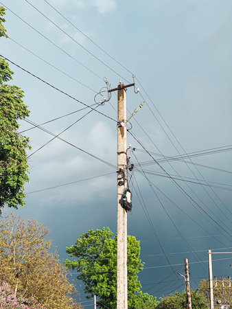 Electrician is scaling a utility pole to conduct maintenance on power lines, surrounded by lush greenery and an ominous stormy sky, showcasing the importance of electrical infrastructureの写真素材