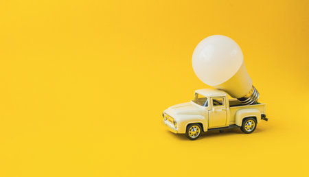 A vintage toy truck features a large light bulb, representing innovation in sustainability, placed on a bright yellow background, evoking creativity and ecological consciousnessの写真素材