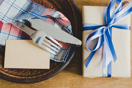 Festive Table Setting. Cutlery with Empty Tag and Present on Linen Napkin on Rustic Wooden Background. Father's Day Concept. Copy Space. Selective Focus.の写真素材