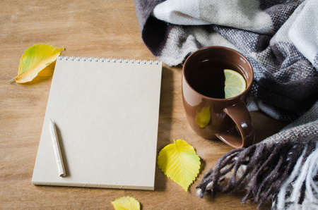 A cup of hot tea with lemon and empty notebook for sketch, plaid on a rustic table, yellow leaves. Cozy autumnal mood, warm autumn.の写真素材