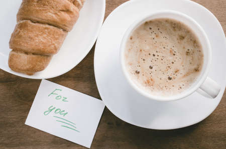 Cappuccino or coffee latte with foam in white cup and croissant on wooden table. Inscription For you.の写真素材