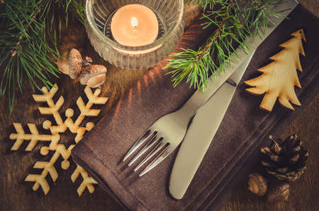 Vintage or rustic Christmas table setting with candle and wooden decorations. Cutlery on linen napkin on rustic wooden background - country style. Selective focus.の写真素材