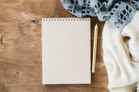 Flat lay of blank brown notebook with knitted blanket on wooden table. Cozy autumn or winter mood....