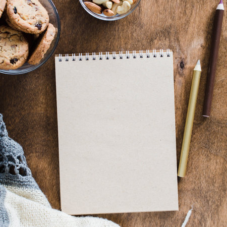 Flat lay of blank brown notebook with knitted blanket and snacks on wooden table. Cozy autumn or...
