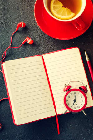 Blank red notebook with pen, headphones and cup of tea on dark table. Business still life, office or education concept. Top view of working desk.の写真素材