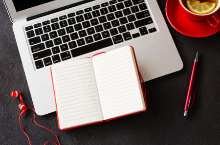 Blank red notebook with pen, computer laptop, headphones and cup of tea on dark table. Business still life, office or education concept. Top view of working desk.の写真素材