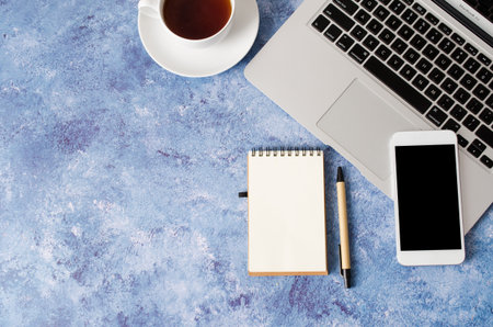 White smartphone with black blank screen on office desk with laptop, empty notebook and cup of tea....