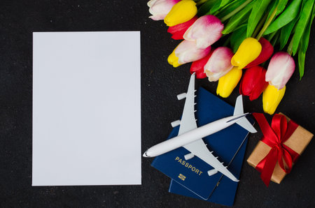 Holiday Travel Planning. Travel concept, mockup. Airplane model, passports, blank paper, bouquet of tulips and gift box on dark background. Top view or flat lay. Copy Space. Mock upの写真素材