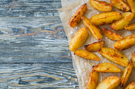 Golden spicy potato wedges fried or oven baked on wooden table. Delicious savoury crisp snack in...