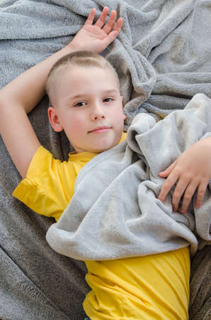 Happy blond boy lying on soft gray blanket at home. Child looking camera. Rest in daytime. Stay at home. Isolation. Quarantine. Top viewの写真素材