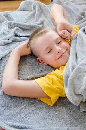 Happy blond boy wakes up at home. Child in yellow T-shirt resting lying on soft gray blanket. Rest in daytime. Stay at home. Isolation. Quarantine. Top viewの写真素材