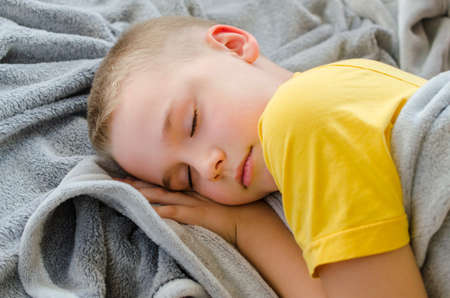 Blond boy sleeps at home. Child in yellow T-shirt resting lying on soft gray blanket. Rest in daytime. Stay at home. Isolation. Quarantine. Top viewの写真素材