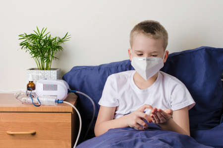 Sick boy in medical protective mask using hand sanitizer. Child sits on bed. Coronavirus Outbreak Conceptの写真素材