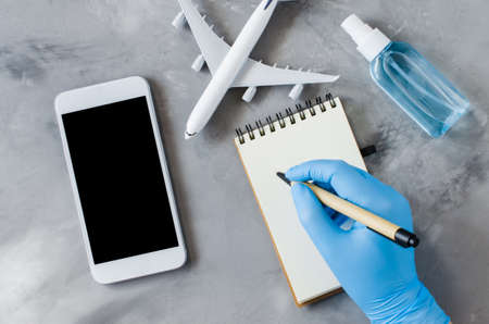 Travel planning concept, coronavirus and quarantine. Hands in disposable gloves hold smartphone and write in notebook. Mock up with copy space. Airplane model, face mask and hand sanitizer spray.の写真素材
