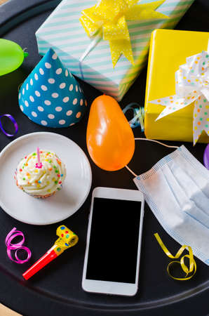 Birthday online or party. Quarantine birthday in isolation. Smartphone, birthday cupcake, medicine mask, gifts and holiday accessories. Social distance. Mock upの写真素材