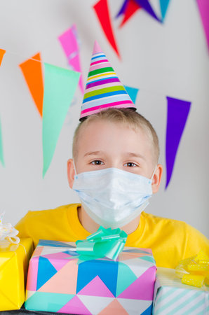 Happy boy in medicine face mask and festive cap with gifts in hand celebrates birthday. Quarantine...