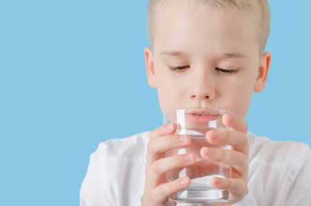 Cute child drinking clean water from glass. Blond little boy with water glass isolated on blue...