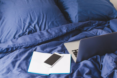 Laptop, smartphone and notebook on the bed in morning time. Bedroom, work from home concept.