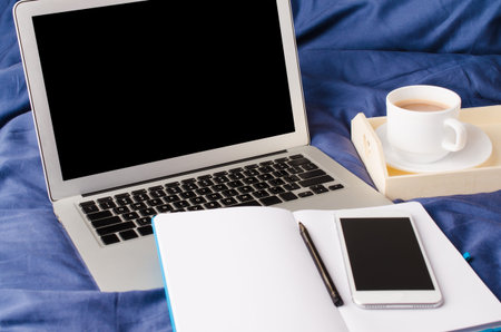 Laptop and smartphone, coffee cup and notebook on the bed in morning time. Mock up. Bedroom, work...