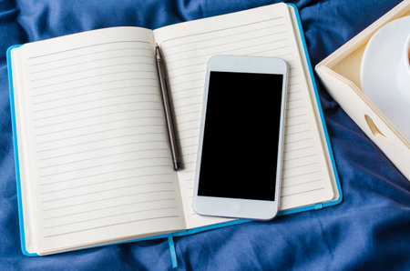 Blank smartphone screen, notebook and cup of coffee on a tray on the bed. Mock up.