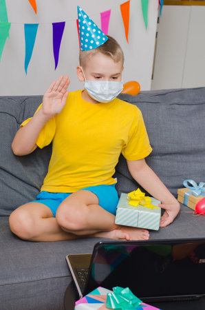 Happy boy in medicine face mask and festive cap with gift celebrates birthday by video call to...