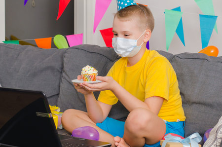 Sad boy in medicine face mask with holiday cupcake celebrates birthday by video call to laptop....
