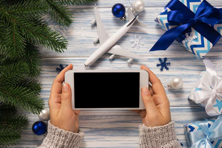 Traveling as gift. Female hands holding a smartphone for travel planning. Christmas background with model of plane and gift boxes. Mockup.の写真素材